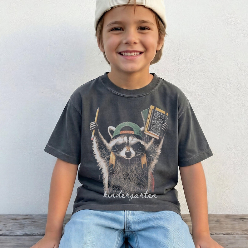 May include: A charcoal grey short-sleeve t-shirt featuring a graphic of a raccoon wearing a green baseball cap and sunglasses, holding a pencil and a notebook. The word kindergarten is printed in white script at the bottom of the graphic.