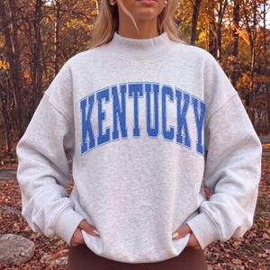 May include: A light gray heathered crewneck sweatshirt featuring the word KENTUCKY printed in a large, arched, distressed blue font across the chest. The garment has long sleeves with ribbed cuffs and a ribbed mock neck collar design.