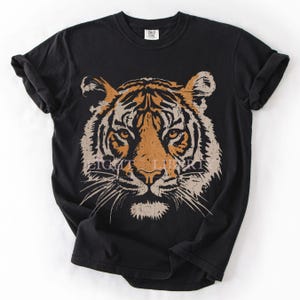 May include: A black short-sleeved t-shirt featuring a large, centred graphic of a tiger face in shades of orange, tan, and cream. The tiger illustration has a distressed, textured appearance. The shirt has a crew neckline and rolled sleeves.