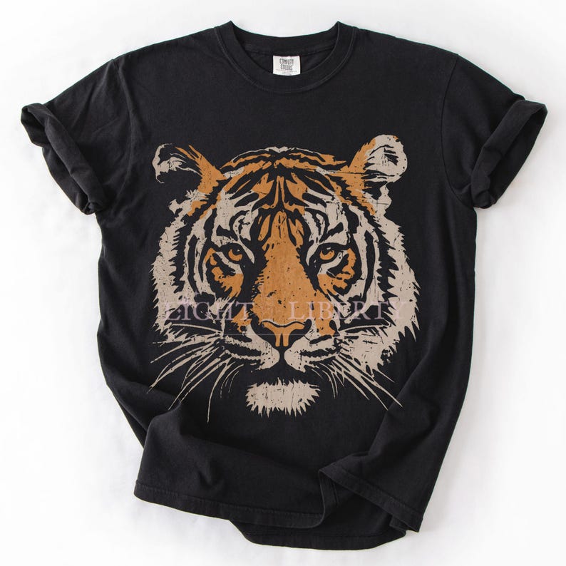 May include: A black short-sleeved t-shirt featuring a large, centred graphic of a tiger face in shades of orange, tan, and cream. The tiger illustration has a distressed, textured appearance. The shirt has a crew neckline and rolled sleeves.