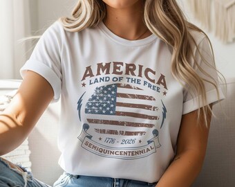 May include: White t-shirt with a distressed graphic of the American flag. The design includes the text "AMERICA LAND OF THE FREE" and "1776-2026 SEMIQUICENTENNIAL" in brown and blue. The shirt has rolled-up sleeves and is worn with blue jeans.