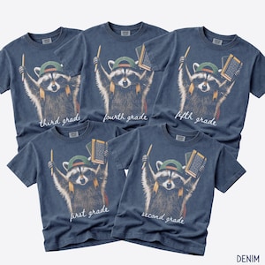 May include: Five denim blue t-shirts featuring a graphic of a raccoon wearing a green baseball cap and sunglasses, holding a pencil and a notebook. Each shirt displays a different grade level in white script: first, second, third, fourth, and fifth grade.