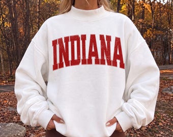 May include: A white long-sleeve sweatshirt featuring a mock neck collar and the word INDIANA printed across the chest in a distressed red collegiate-style font. The garment has a relaxed fit with ribbed cuffs and a ribbed hemline.