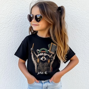 May include: A black short-sleeved t-shirt featuring a graphic of a raccoon wearing a green baseball cap and sunglasses, holding a pencil and books, with the text pre-k printed below. The shirt is worn with light blue denim jeans.