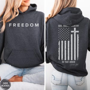May include: Dark heather gray hoodie with the word "FREEDOM" printed across the chest. The back features an American flag design with a cross and the text "GOD, FAMILY, COUNTRY IN THAT ORDER".