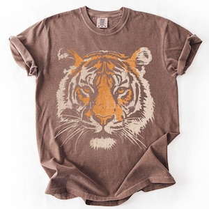 May include: A brown short-sleeved t-shirt featuring a large, distressed graphic print of a tiger face on the front. The tiger design is rendered in shades of orange and cream, showcasing detailed facial features, whiskers, and stripes.