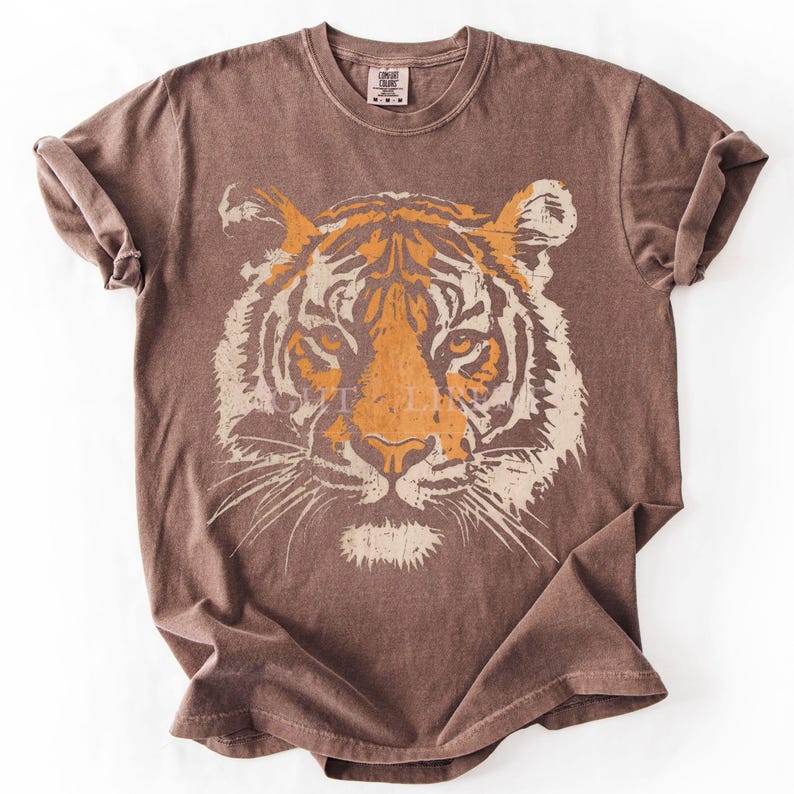 May include: A brown short-sleeved t-shirt featuring a large, distressed graphic print of a tiger face on the front. The tiger design is rendered in shades of orange and cream, showcasing detailed facial features, whiskers, and stripes.