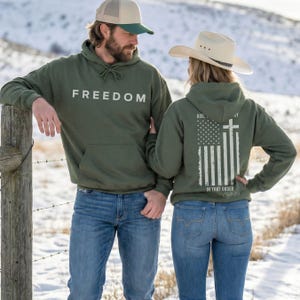 May include: Two people wearing olive green hoodies and blue jeans. The hoodie on the left has the word "FREEDOM" printed in white. The hoodie on the right features an American flag design with a cross and the text "GOD, COUNTRY, IN THAT ORDER".
