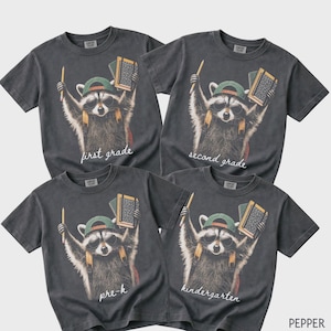 May include: Four charcoal grey short-sleeve t-shirts featuring a graphic of a raccoon wearing sunglasses, a green cap, and a backpack. The raccoon holds a pencil and a notebook. Text on the shirts reads first grade, second grade, pre-k, and kindergarten.
