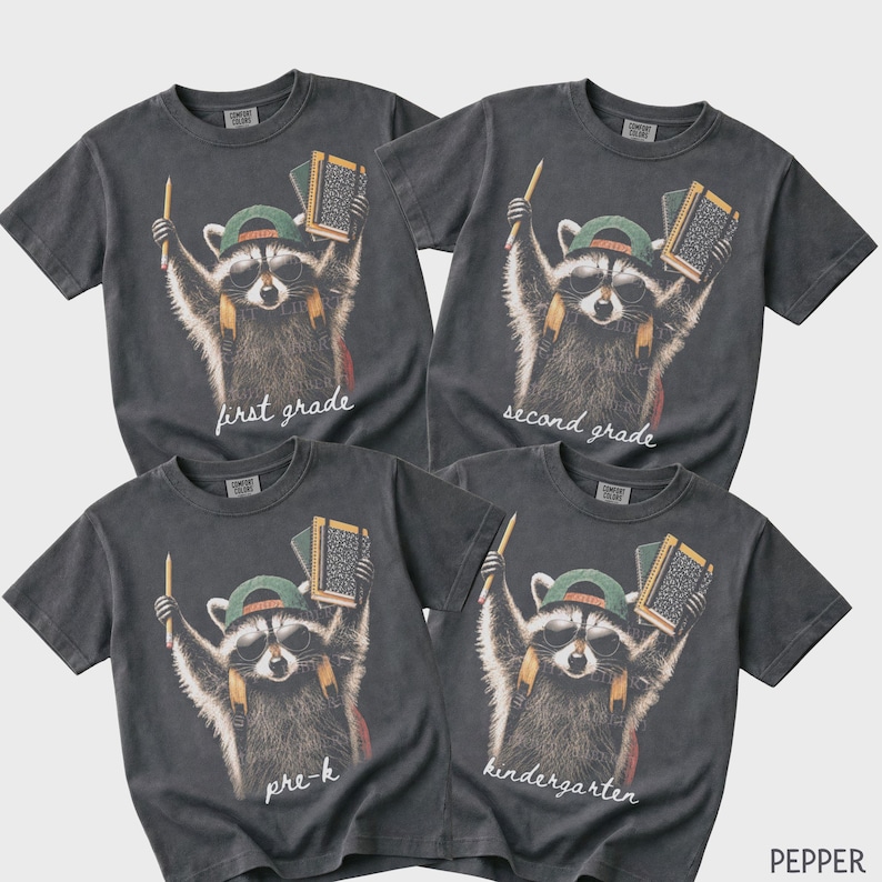 May include: Four charcoal grey short-sleeve t-shirts featuring a graphic of a raccoon wearing sunglasses, a green cap, and a backpack. The raccoon holds a pencil and a notebook. Text on the shirts reads first grade, second grade, pre-k, and kindergarten.