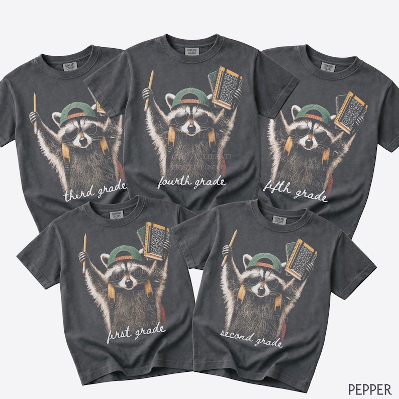 May include: Five charcoal grey t-shirts featuring a graphic of a raccoon wearing sunglasses and a green baseball cap, holding a pencil and a book. Each shirt displays a different grade level text: first grade, second grade, third grade, fourth grade, fifth grade.