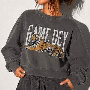 May include: A charcoal gray crewneck sweatshirt featuring a graphic of a resting tiger in orange and black tones. The text "GAME DEY" is printed in large, distressed white capital letters above the tiger illustration. The garment has long sleeves.