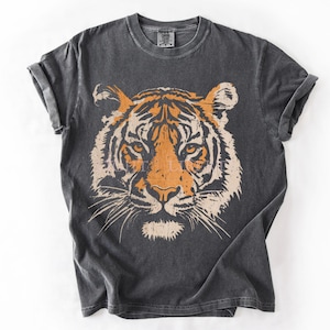 May include: A charcoal grey short-sleeve t-shirt featuring a large, centred graphic of a tiger face in shades of orange and cream. The tiger illustration has a distressed, textured appearance. The shirt has a crew neckline and rolled sleeves.