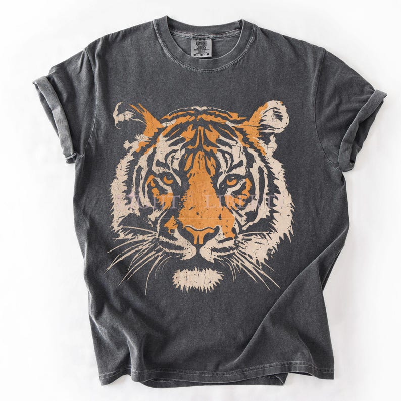 May include: A charcoal grey short-sleeve t-shirt featuring a large, centred graphic of a tiger face in shades of orange and cream. The tiger illustration has a distressed, textured appearance. The shirt has a crew neckline and rolled sleeves.