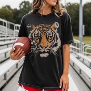 May include: A charcoal grey short-sleeve t-shirt featuring a large, detailed graphic of a tiger face in shades of orange, black, and white. The shirt has a crew neckline and a relaxed fit, showcasing a vintage-inspired, faded fabric texture.