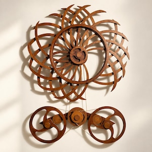 May include: A large brown wooden wall sculpture featuring a complex circular gear-like design with curved spokes. Below the main circular element, two smaller circular components are connected by thin strings to the central mechanism.