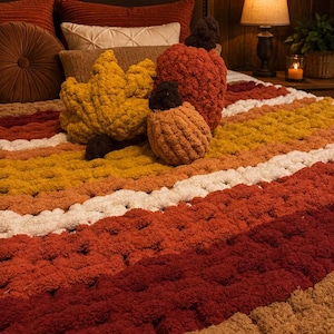 May include: A chunky knit throw blanket featuring horizontal stripes in shades of burgundy, orange, yellow, white, and tan. Resting on the blanket are three plush decorative pillows shaped like a large yellow leaf, a large orange pumpkin, and a small orange pumpkin.