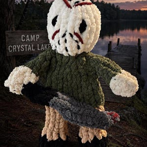 May include: A plush doll featuring a white hockey mask with red markings, a dark green textured sweater, tan pants, and black boots. The doll holds a gray machete with a red tip. A wooden sign in the background reads CAMP CRYSTAL LAKE.