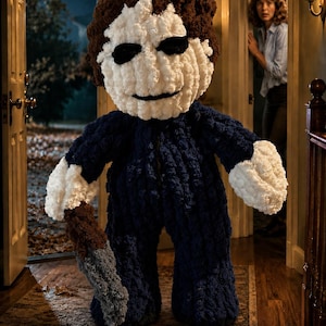 May include: A plush, crocheted figure resembling a horror movie character with a white mask, dark brown hair, and a navy blue jumpsuit. The figure has white hands and holds a gray, textured knife-shaped object in its right hand.