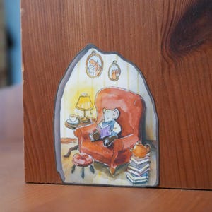 Op de afbeelding: Waterverfillustratie van een muis die leest in een rode fauteuil, met een lamp, een tafeltje en een stapel boeken. De afbeelding is geplaatst in een houten frame, wat een gezellige, sprookjesachtige sfeer creëert.