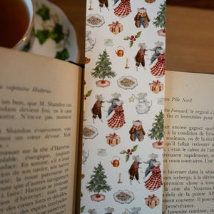 May include: A decorative bookmark with a repeating pattern of dancing mice, Christmas trees, gifts, and teacups. The mice are dressed in festive attire. The bookmark is placed in an open book with text.