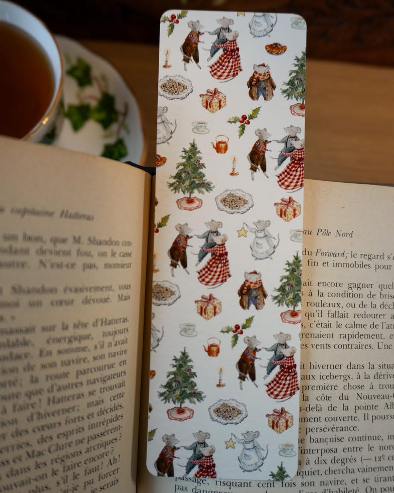 May include: A decorative bookmark with a repeating pattern of dancing mice, Christmas trees, gifts, and teacups. The mice are dressed in festive attire. The bookmark is placed in an open book with text.