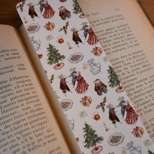 May include: A whimsical bookmark featuring a repeating pattern of dancing mice in festive attire, Christmas trees, gifts, and teacups. The bookmark is set against the backdrop of an open book, enhancing its literary theme.