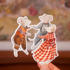 May include: A die-cut sticker featuring three anthropomorphic mice dancing together. One mouse wears an orange vest and plaid shirt, another wears a grey shirt with suspenders and a red bowtie, and the third wears a red and white checkered dress.
