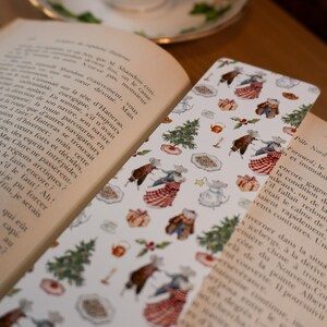 May include: A white bookmark with a festive pattern featuring dancing mice, Christmas trees, and gifts. The bookmark is placed between the pages of an open book, with a teacup and saucer in the background.