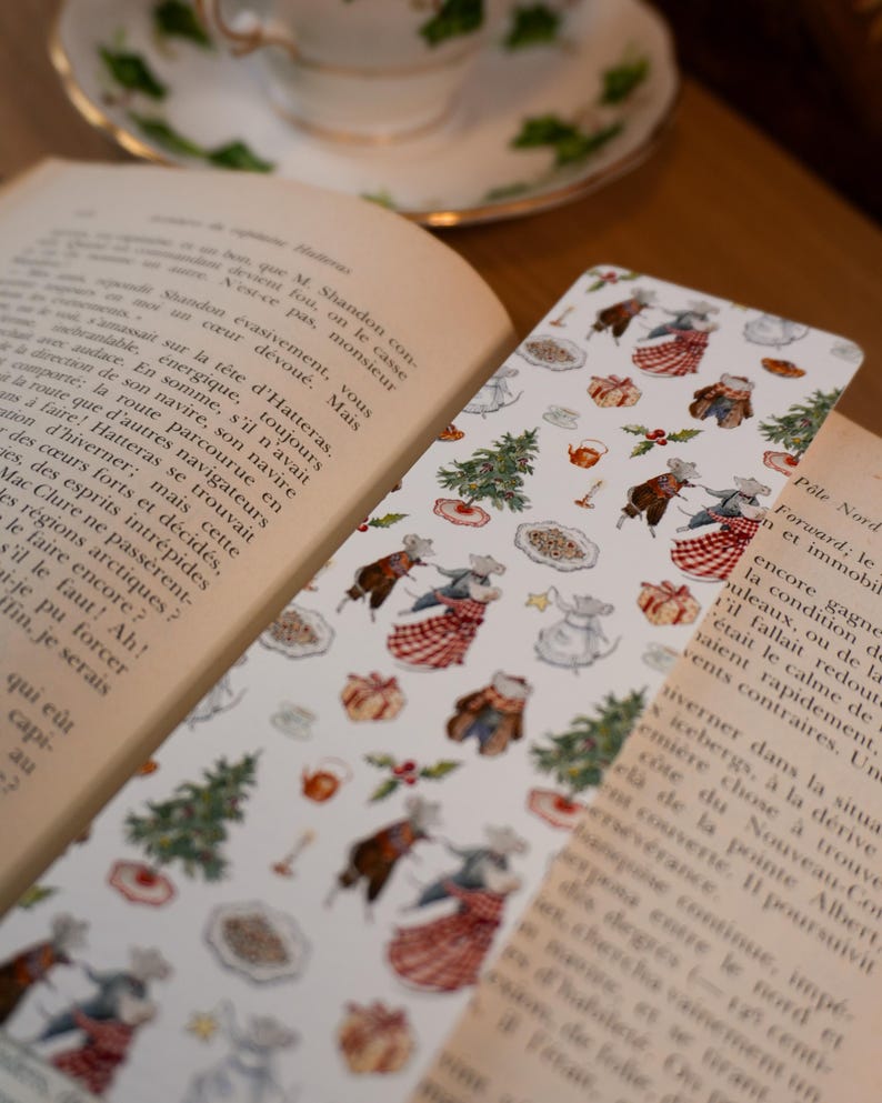 May include: A white bookmark with a festive pattern featuring dancing mice, Christmas trees, and gifts. The bookmark is placed between the pages of an open book, with a teacup and saucer in the background.