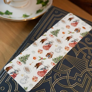 May include: A whimsical bookmark featuring a repeating pattern of festive mice, Christmas trees, and gifts. The bookmark rests on a dark blue book with gold embossed detailing, next to a teacup with a green floral design.