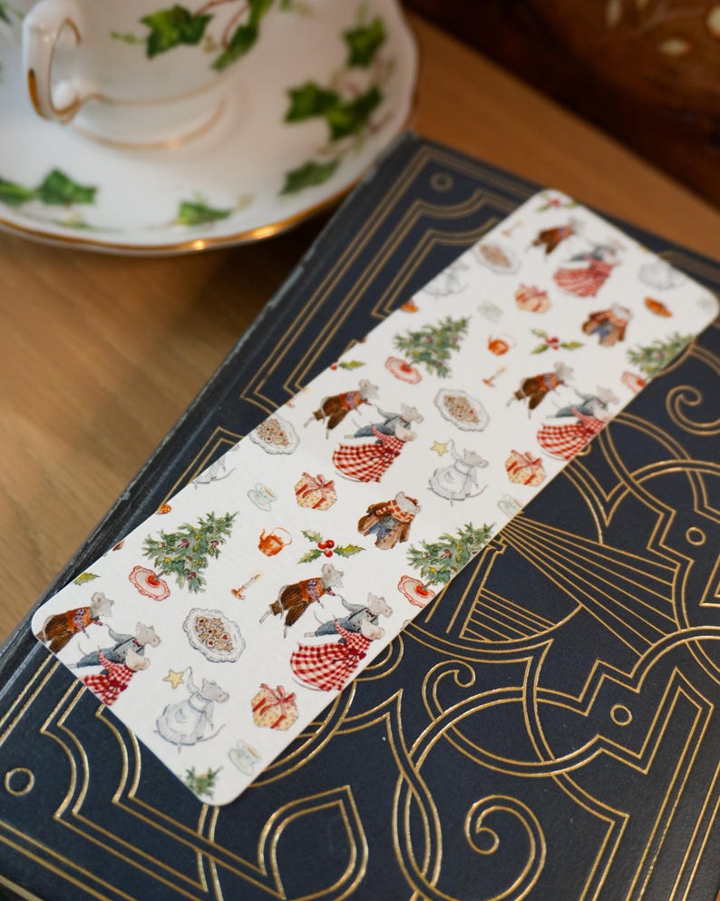 May include: A whimsical bookmark featuring a repeating pattern of festive mice, Christmas trees, and gifts. The bookmark rests on a dark blue book with gold embossed detailing, next to a teacup with a green floral design.