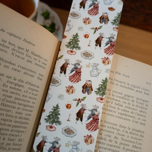 Puede incluir: Un marcapáginas blanco con un patrón repetido de ratones bailando, árboles de Navidad, regalos y platos de comida. El marcapáginas está en un libro abierto con texto en francés. El tema es festivo y caprichoso.