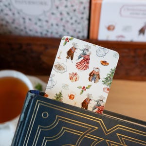 May include: A decorative bookmark featuring a whimsical illustration of mice in festive attire, set against a white background with Christmas-themed elements. The bookmark is inserted into a dark blue book with gold accents.