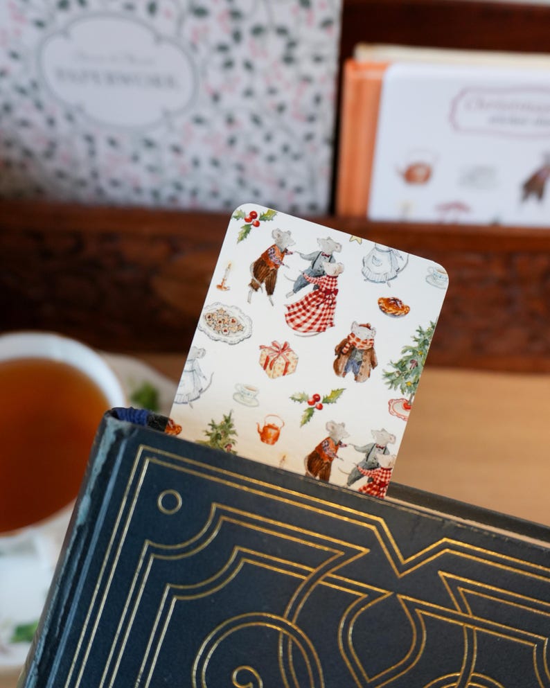 May include: A decorative bookmark featuring a whimsical illustration of mice in festive attire, set against a white background with Christmas-themed elements. The bookmark is inserted into a dark blue book with gold accents.
