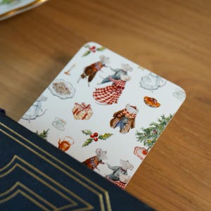 May include: A decorative bookmark featuring a whimsical illustration of mice in festive attire, surrounded by Christmas-themed elements like gifts, holly, and a Christmas tree. The bookmark is inserted into a navy blue book with gold accents.
