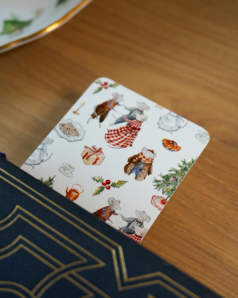 May include: A decorative bookmark featuring a whimsical illustration of mice in festive attire, surrounded by Christmas-themed elements like gifts, holly, and a Christmas tree. The bookmark is inserted into a navy blue book with gold accents.