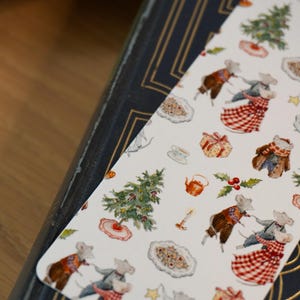 May include: A whimsical bookmark featuring a repeating pattern of dancing mice, Christmas trees, gifts, and cookies. The bookmark is white with a decorative border and is resting on a dark blue book with gold accents. The overall theme is festive and charming.