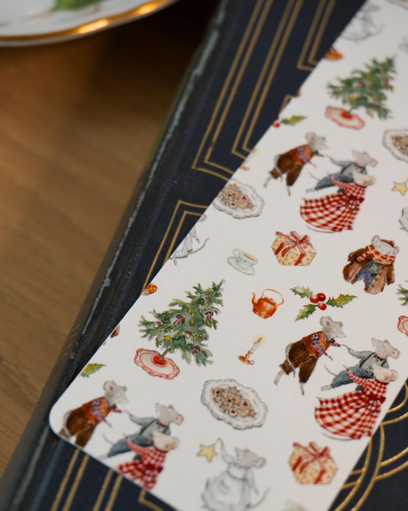 May include: A whimsical bookmark featuring a repeating pattern of dancing mice, Christmas trees, gifts, and cookies. The bookmark is white with a decorative border and is resting on a dark blue book with gold accents. The overall theme is festive and charming.