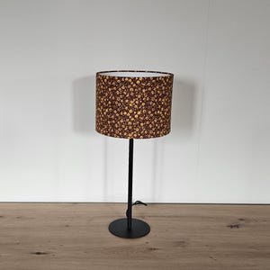 Op de afbeelding: Een zwarte metalen tafellamp met een cilindrische lampenkap. De lampenkap heeft een bruin bloemenpatroon met gele bloemen. De lamp heeft een ronde voet en een rechte steel.