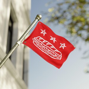 May include: A red flag with a white illustration of a sandwich and three stars. The flag is mounted on a silver pole and is waving in the wind against a blue sky and a building.