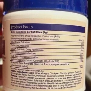 Puede incluir: Un envase blanco y azul con tapa azul, que muestra una etiqueta de datos del producto. La etiqueta enumera los ingredientes activos por masticaci&oacute;n blanda, incluyendo una mezcla patentada de probi&oacute;ticos, calostro bovino seco y otros extractos. El envase es sostenido por una persona.