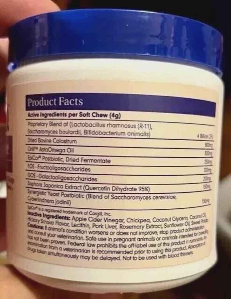 Puede incluir: Un envase blanco y azul con tapa azul, que muestra una etiqueta de datos del producto. La etiqueta enumera los ingredientes activos por masticaci&oacute;n blanda, incluyendo una mezcla patentada de probi&oacute;ticos, calostro bovino seco y otros extractos. El envase es sostenido por una persona.