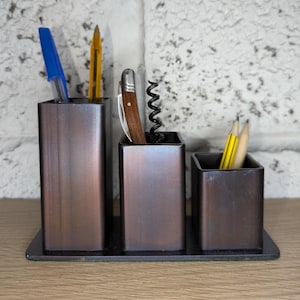 May include: Three bronze metal desk tidies on a black base. They contain a blue pen, yellow pencil, corkscrew, and pencils. The background is a white brick wall.