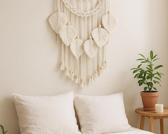 Puede incluir: Colgante de pared de macramé color crema con una clavija de madera y formas de hojas decorativas. El arte de la pared presenta detalles anudados, cuentas de madera y borlas. Esta decoración de estilo bohemio añade un elemento natural y texturizado.
