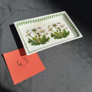 May include: A rectangular white ceramic tray with a floral design featuring white daisies with yellow centres and green leaves. The tray has a green border with a leaf pattern. A red sticky note with the number 4.3 is in the foreground.
