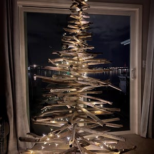 May include: A rustic, handmade driftwood Christmas tree illuminated with warm white fairy lights. The tree is constructed from various pieces of weathered wood, arranged in a conical shape. The tree is displayed in front of a window with a night view.