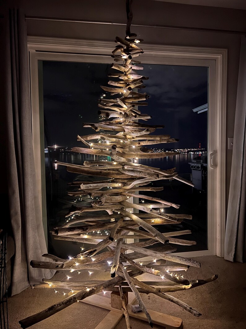 May include: A rustic, handmade driftwood Christmas tree illuminated with warm white fairy lights. The tree is constructed from various pieces of weathered wood, arranged in a conical shape. The tree is displayed in front of a window with a night view.