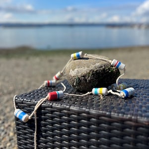 May include: A string of decorative buoys with red, white, blue, and yellow stripes, resting on a dark brown woven surface. The buoys are connected by a white rope and are near a rock covered in moss. The background shows a beach and ocean.