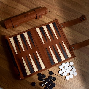 May include: A brown backgammon set with black, white, and tan triangular points. The set includes black and white game pieces, dice, and a brown leather carrying case. The game is open on a wooden surface.